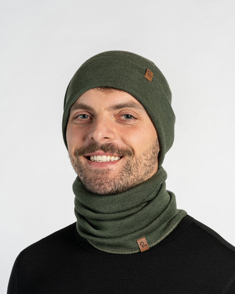 A smiling man with short dark hair and a beard, wearing a dark green menique knit beanie with a small brown tag and a matching dark green knit neck warmer, looks directly at the camera. He is wearing a dark long-sleeved shirt. The background is plain white.