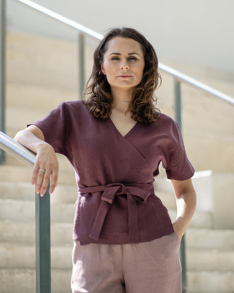 A woman with brown hair, wearing a dark purple wrap top and light mauve trousers, smiles while posing on indoor concrete stairs.