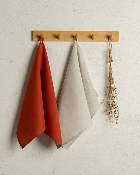 Two menique linen kitchen dish towels hanging on a wooden rack on a wall. The colors of the towels are cinnamon red and natural. 