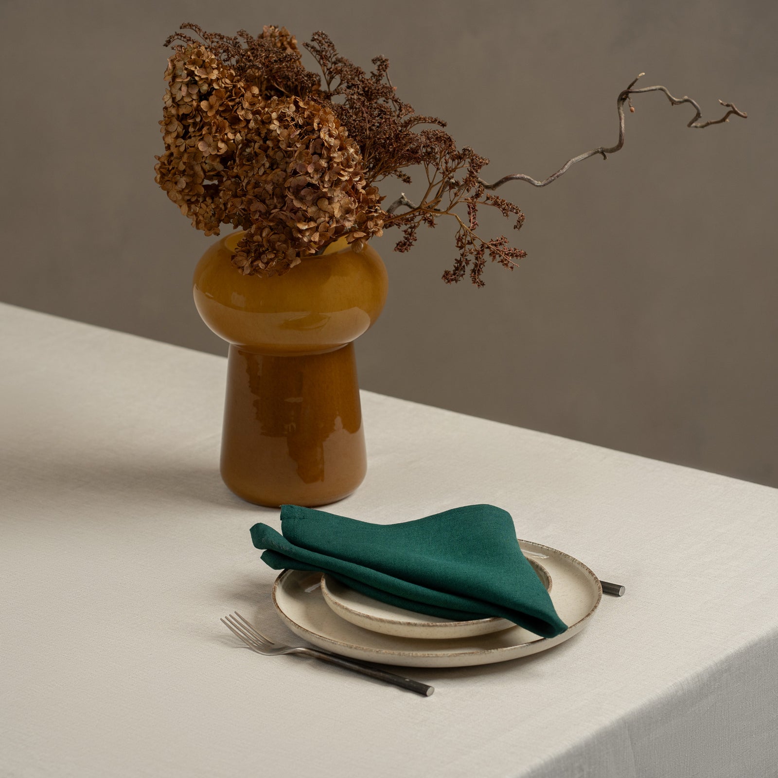 A tabletop covered with a linen tablecloth, with a vase and a single plate set on top of it. By the sides of the plates there are cutlery items, and there is a linen napkin in cobalt blue color on top.