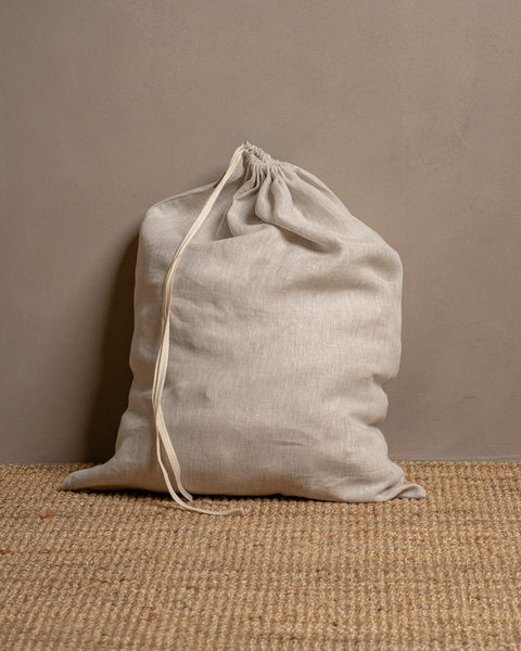 A closed, natural color linen storage bag sits on a textured jute rug against a warm brown wall. The drawstrings are pulled closed at the top.