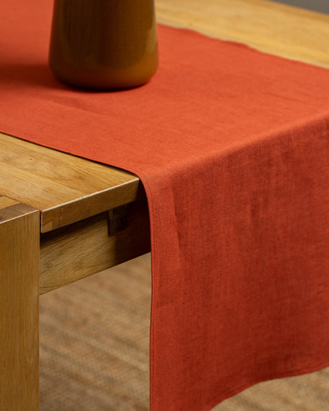 The cinnamon red linen table runner on top of a wooden table, captured in a close up picture from the corner of the table. 