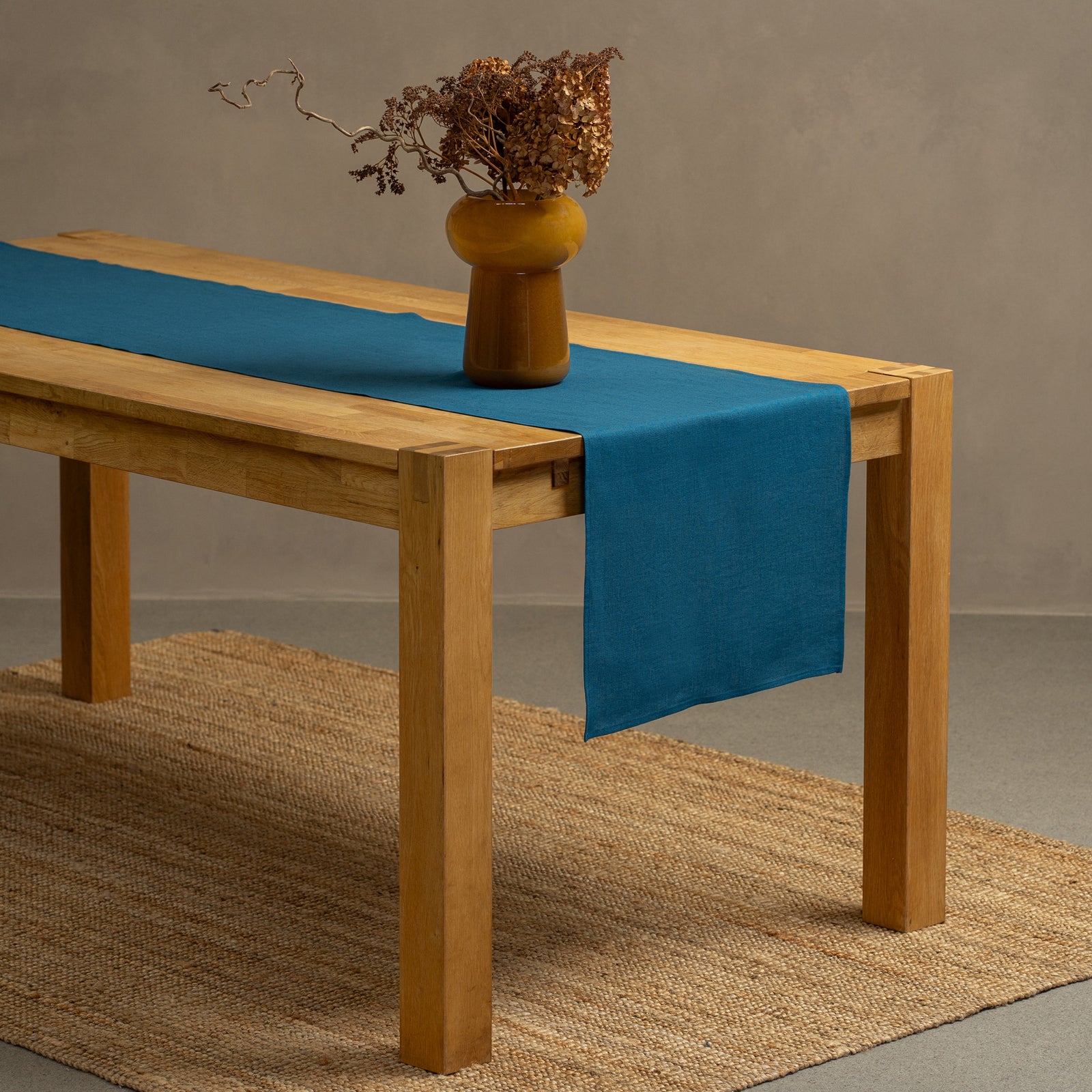 A wooden table positioned on top of a carpet. There is a dark green linen table runner and a vase on top.