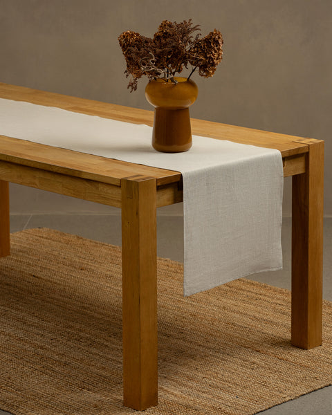 A wooden table positioned on top of a carpet. There is a natural linen table runner and a vase on top.