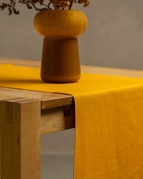 The spicy yellow linen table runner on top of a wooden table, captured in a close up picture from the corner of the table. 