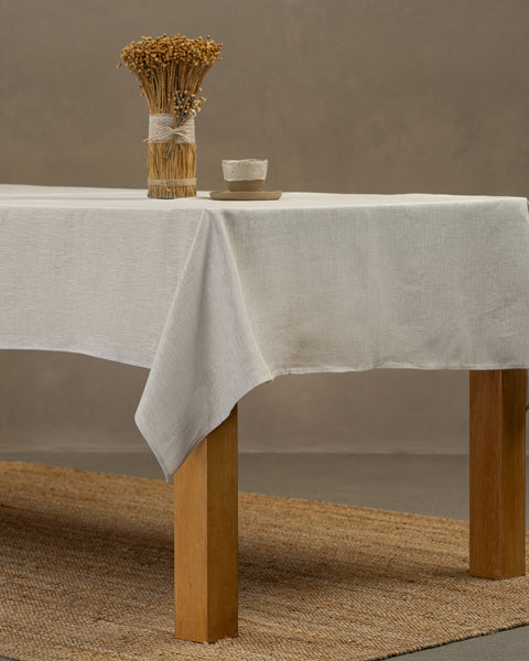 The menique linen tablecloth in pure white color, draping over a wooden table, with a vase and a clay cup and plate set on top of it.