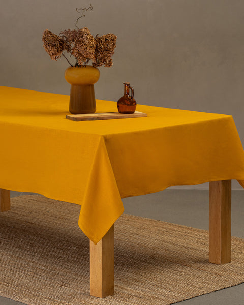The menique linen tablecloth in spicy yellow color, draping over a wooden table, with a vase, wooden board and a glass bottle on top of it.