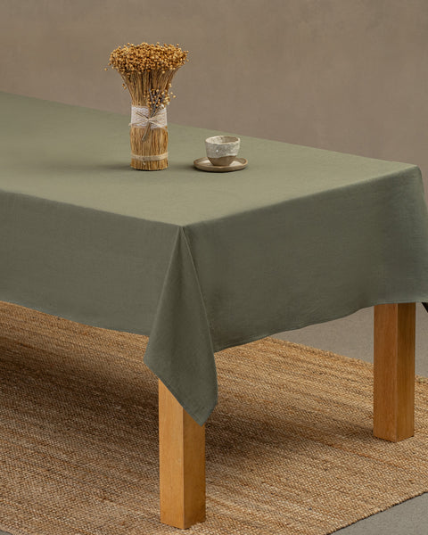 The menique linen tablecloth in stone green color, draping over a wooden table, with a vase on top of it, and a clay cup and plate by the side.