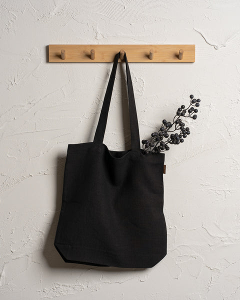Menique reusable pure black color linen tote bag hangs from a wooden rack with four pegs mounted on a textured off-white wall.