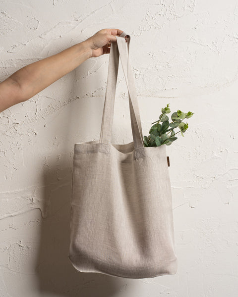 A fair-skinned arm and hand hold up a natural color reusable linen tote bag against a textured off-white wall. Green leafy stems are visible inside the bag.