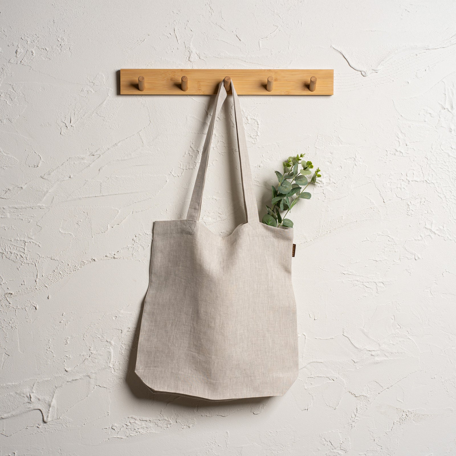 Menique natural color linen tote bag with long handles hangs from a branch of a light-colored tree. In the blurred background, there is a body of water reflecting the sky, surrounded by green grass and trees under a partly cloudy sky.