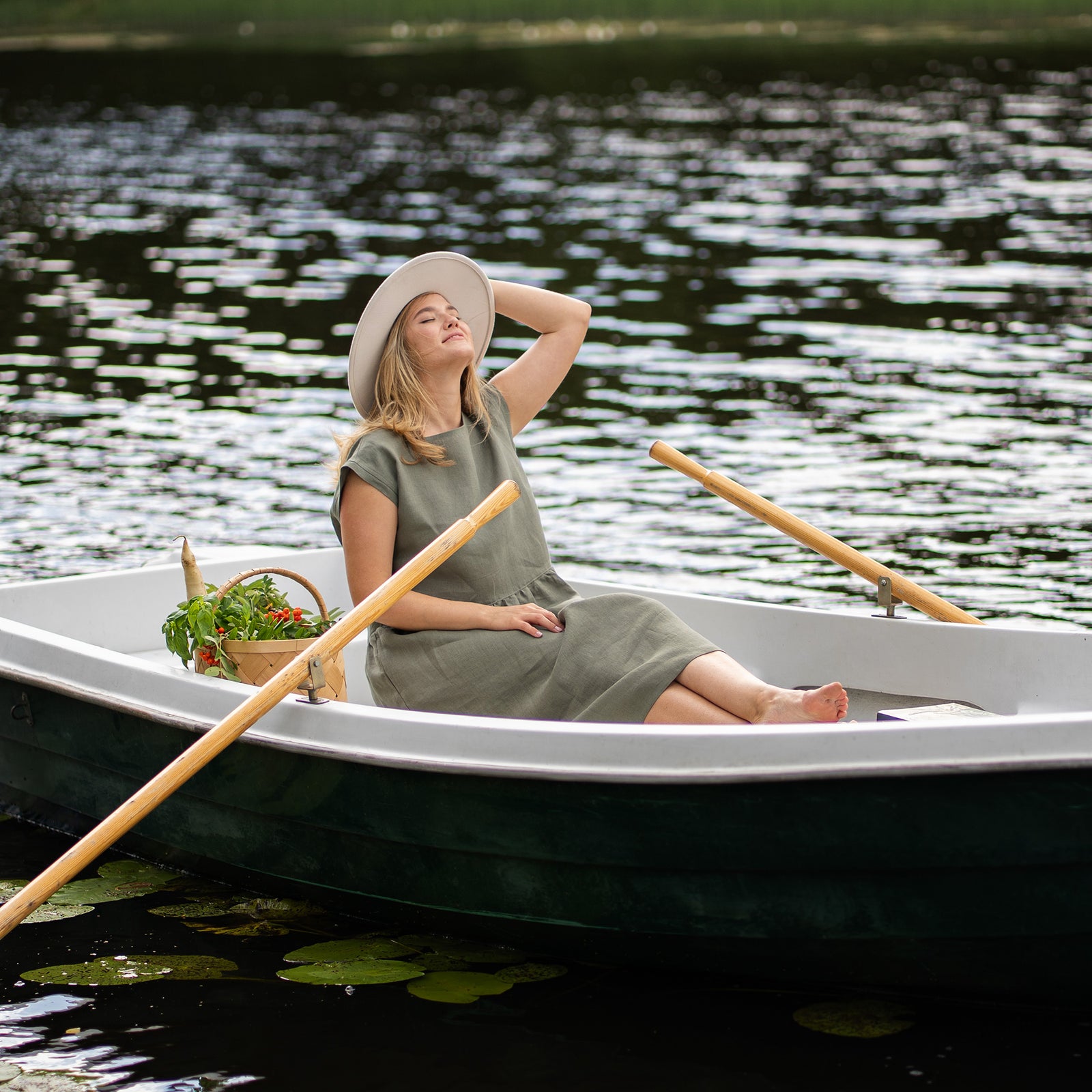 A woman sits in a white rowboat holding a vibrant bouquet of sunflowers, her stone green linen dress complementing the natural outdoor setting. She looks directly at the camera while gently touching her hair, with a light-colored hat resting on the boat's seat nearby.