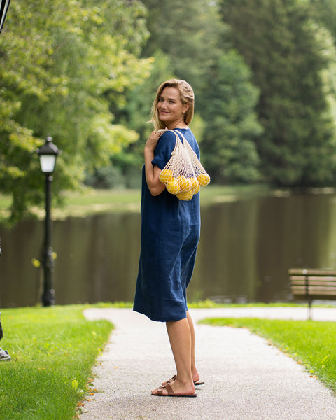 A smiling woman stands on a park pathway, looking back over her shoulder at the camera while wearing a storm blue linen dress and brown sandals. She casually carries a string bag full of lemons over her shoulder, with a tranquil lake and trees in the background.