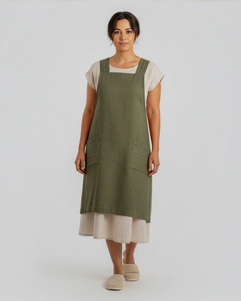A woman standing in a studio setting wearing a stone green linen Japanese apron featuring two front pockets, layered over a cream dress and soft slippers.