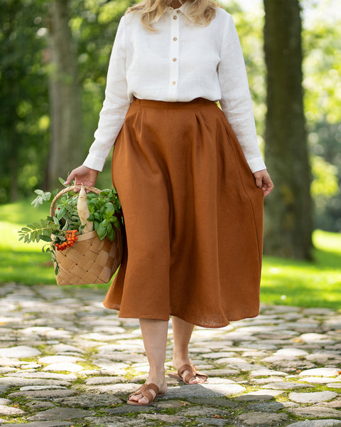 A woman walks gracefully along a dappled cobblestone path, carrying a woven basket overflowing with fresh greens, roots, and bright red berries. She is dressed in a pure white button-down linen shirt tucked into a long, beautifully draped almond brown maxi skirt.