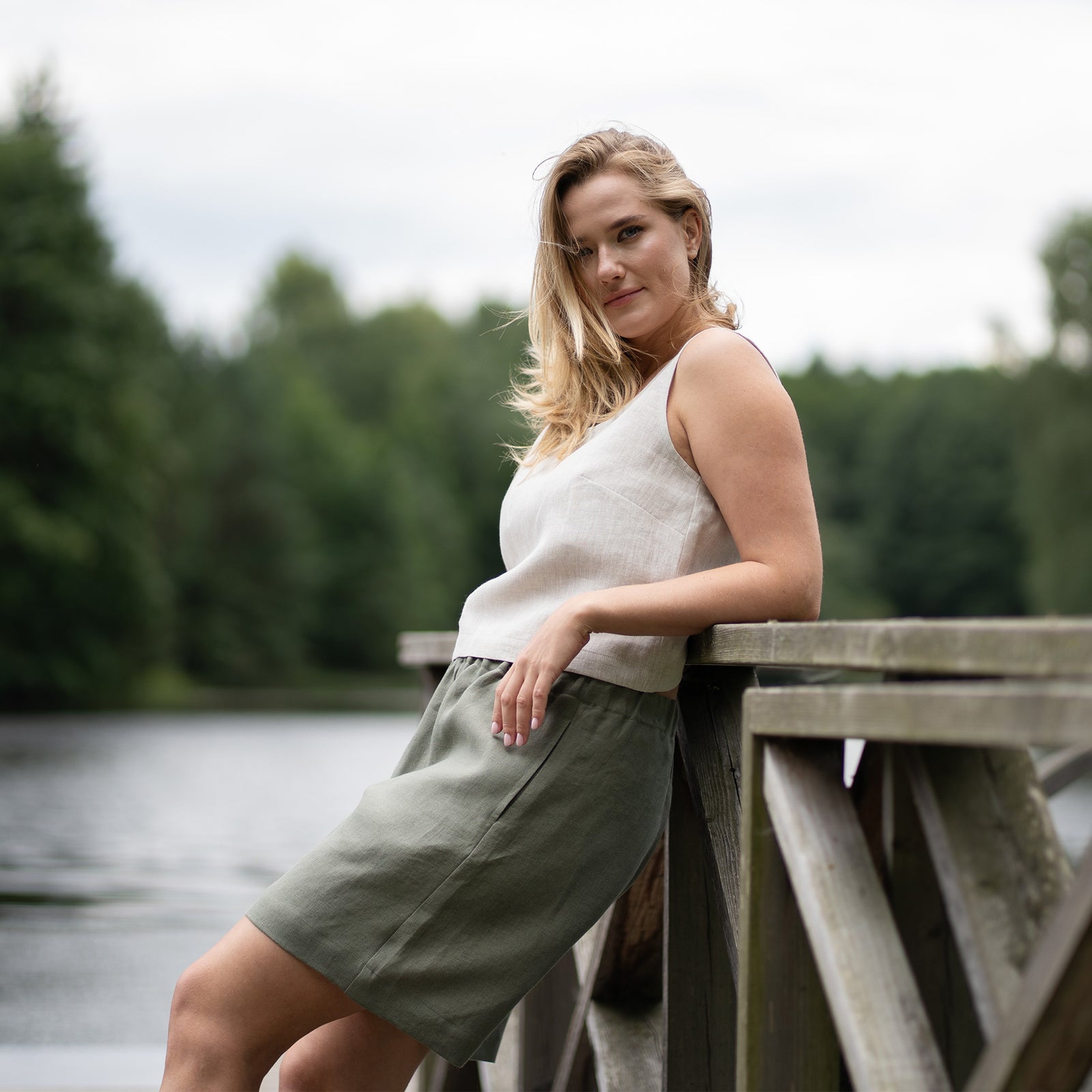 A barefoot woman strolls down a sun-dappled dirt path through a lush green forest. She is wearing a natural-colored linen crop top paired with comfortable stone green linen Bermuda shorts.