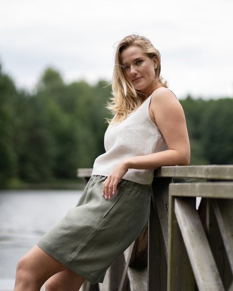 A woman leans casually against a wooden pier railing, wearing a sleeveless natural linen top and stone green bermuda shorts. The wind catches her hair as she looks toward the camera, with a calm lake and forest setting the scene behind her.