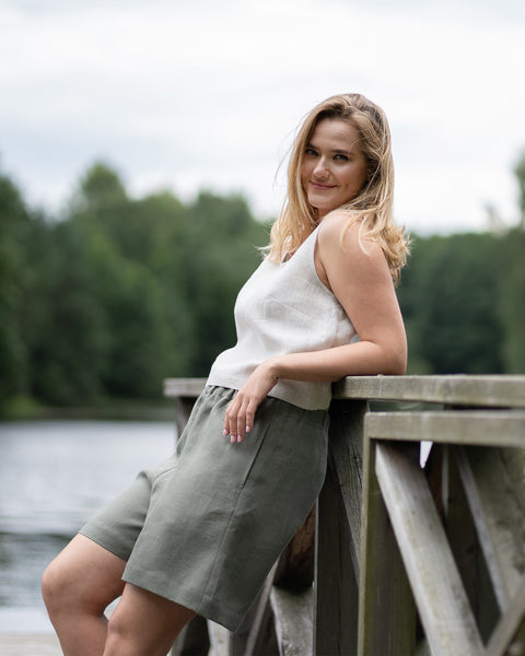 A barefoot woman strolls down a sun-dappled dirt path through a lush green forest. She is wearing a natural-colored linen crop top paired with comfortable stone green linen Bermuda shorts.