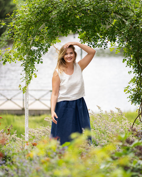 A joyful woman walks through a vine-covered garden archway, touching her hair lightly as the wind blows. She is dressed in a pure white sleeveless linen top paired with a dark blue knee-length skirt.