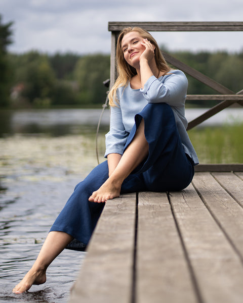 A woman wearing a light blue top and dark blue pants sits comfortably on a wooden dock. She is leaning slightly back with her eyes closed and a peaceful smile, dangling one foot near the water.