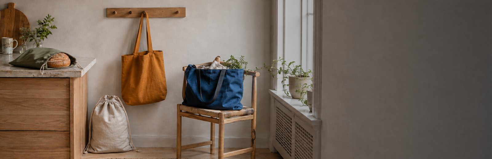 A serene, minimalist interior featuring a collection of reusable fabric bags. A mustard yellow tote bag hangs from a wooden peg rail on a neutral wall. Below it, a beige drawstring bag sits on the wooden floor. To the right, a large blue canvas tote rests on a rustic wooden chair. On the left, a wooden cabinet with a stone countertop holds a dark green fabric bag containing a round loaf of bread, alongside a wooden cutting board and a small vase of greenery.