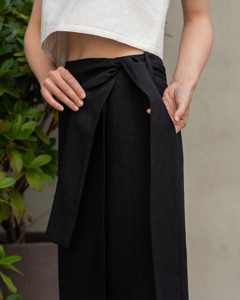 A person is shown from the waist down, adjusting a black wrap skirt. They are wearing a light-colored, possibly white, cropped top that exposes their midriff. The skirt has a prominent tie detail at the waist. In the background, green foliage is visible.