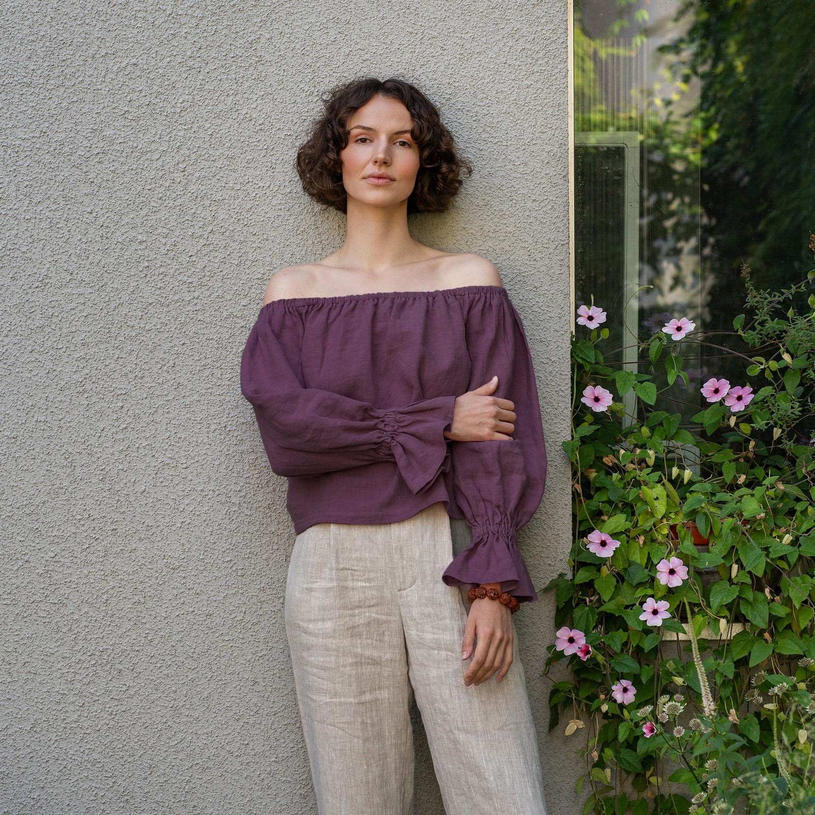 A woman with short, curly brown hair looks directly at the camera, standing in front of a green ivy-covered wall. She is wearing a pure red off-the-shoulder linen top with voluminous sleeves that gather at the wrists, and light-colored pants.