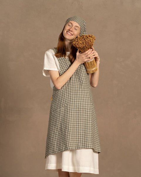 A person with fair skin and long reddish-brown hair stands smiling with closed eyes, gently holding a bundle of dried golden flowers to their chest. They are wearing a white short-sleeved linen shirt under a green check apron and a matching checkered headscarf tied under their chin. The background is a muted warm brown.