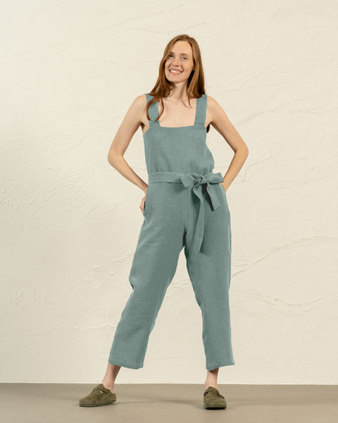 A smiling person with long reddish-brown hair and light skin is standing with their hands on their hips. They are wearing a pinafore jumpsuit with wide straps and a tied belt, along with olive-green slip-on shoes.