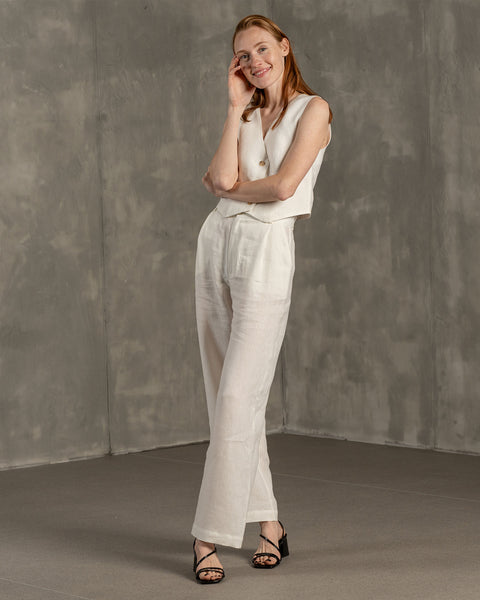 A smiling woman with reddish-brown hair stands in a three-quarter pose, wearing a pure white menique linen vest and wide-leg pants, paired with black open-toe heeled sandals. She has one hand gently touching her cheek and is looking towards the viewer, set against a textured gray wall and floor.