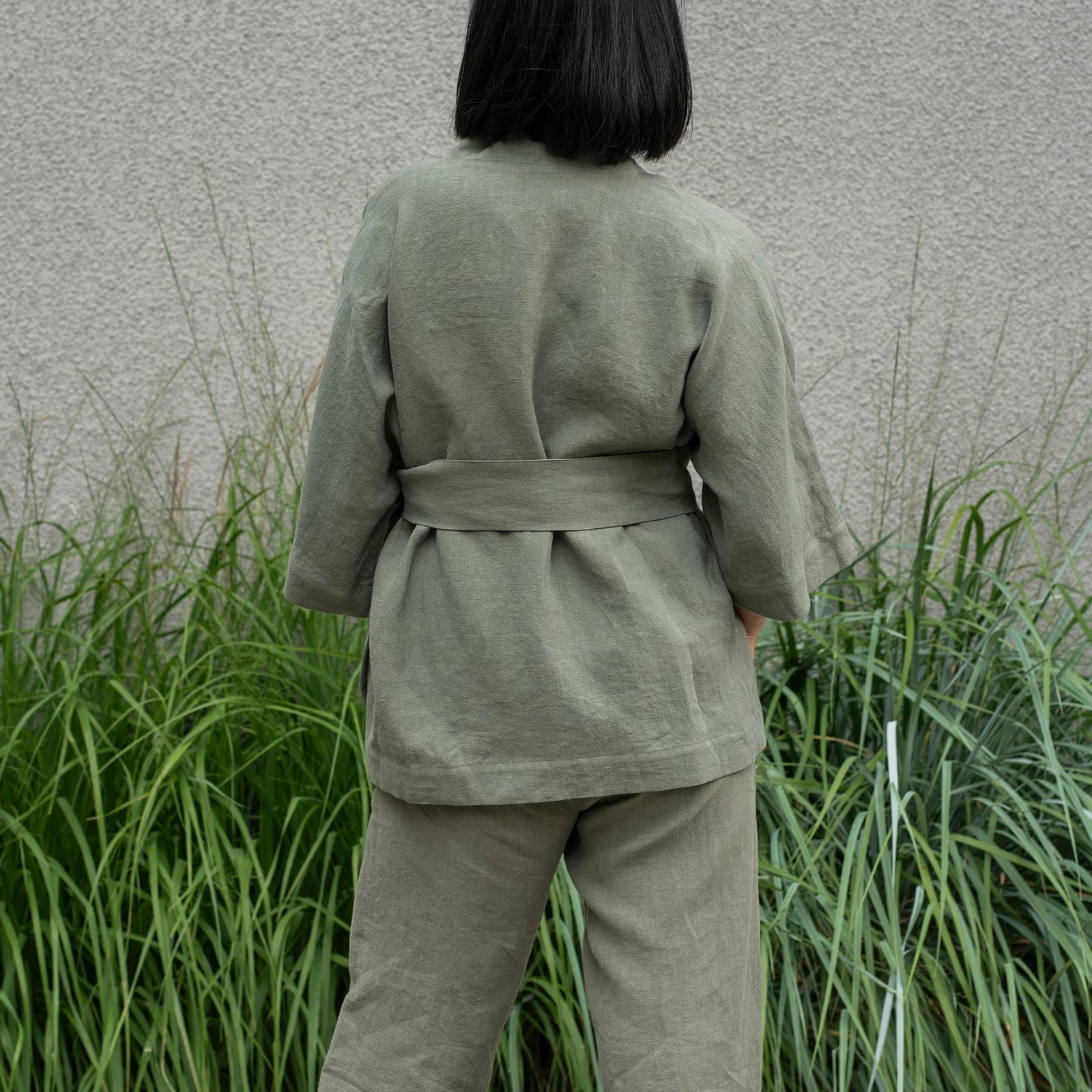 A person stands outdoors, wearing a stone green colored linen kimono-style top and matching wide-leg pants. Their right hand is in the pocket of their pants. To their left, there are tall green plants.