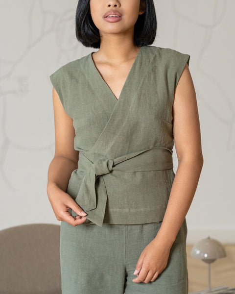 A person with short, dark hair wears a stone green linen wrap top and matching menique pants, tied at the waist, standing indoors.