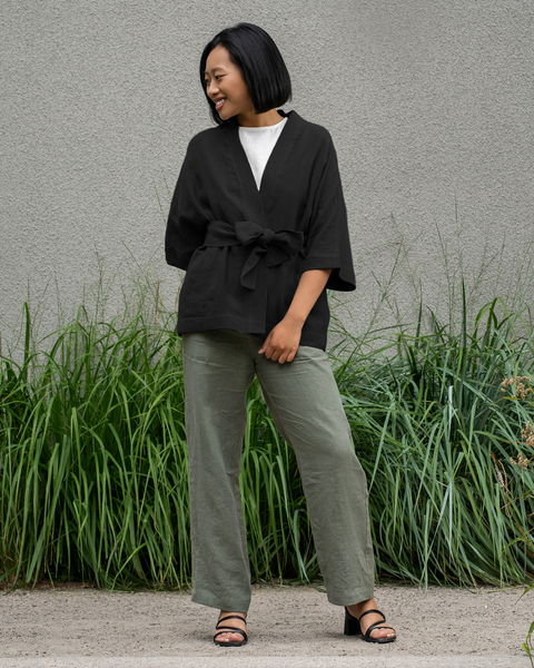 A woman with short dark hair smiles and looks to the right, wearing a pure black linen jacket, stone green wide-leg menique pants and black sandals. She is standing on a paved surface in front of a textured gray wall with tall green plants growing along the base.