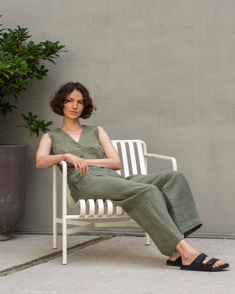 A woman with short curly brown hair is sitting in a white outdoor chair. She is wearing a sleeveless stone green linen vest and matching wide-legged linen pants. She is looking directly at the camera. Behind the chair is a plain gray wall. To the left of the chair is a large brown pot containing a leafy green plant. The ground is light gray concrete.
