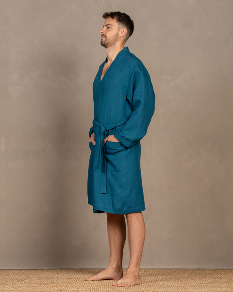A man turned to the side, wearing the cobalt blue menique linen bathrobe. Both of his hands are in the side pockets. The front tie is tied loosely.