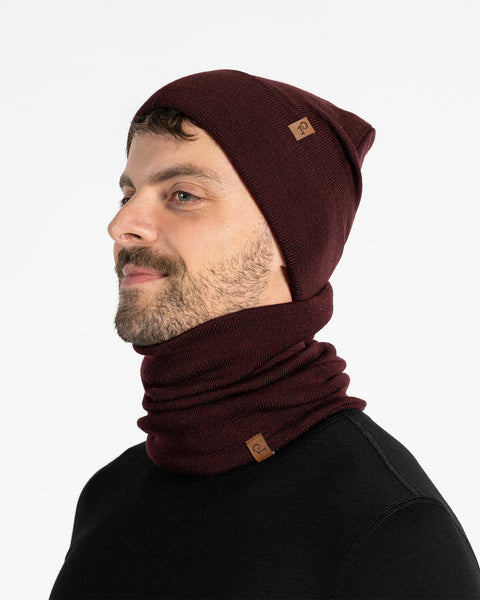A man with a light brown beard is shown from the chest up, looking to the left of the frame. He is wearing a royal cherry knitted beanie and a matching neck gaiter. Both items of clothing have a small brown leather patch with a menique logo on them. The background is a plain white.