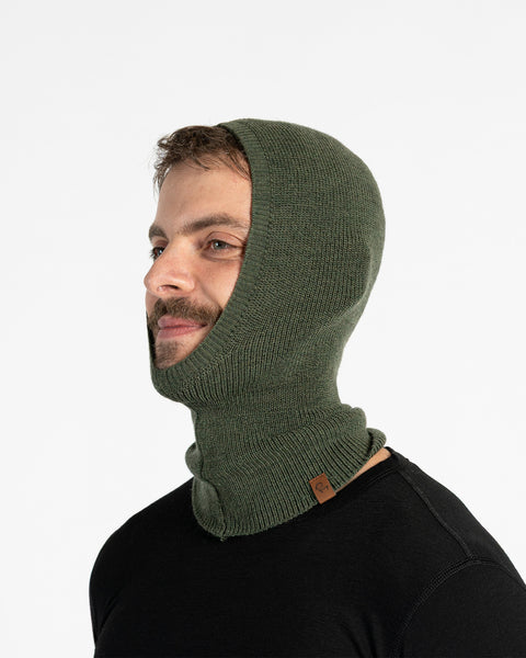 A man with dark hair, a beard, and a mustache is seen from the side, wearing a dark green knitted balaclava that covers his head and neck, with an opening around his face. He is also wearing a dark blue long-sleeved shirt and appears to be looking off to his right with a slight smile.