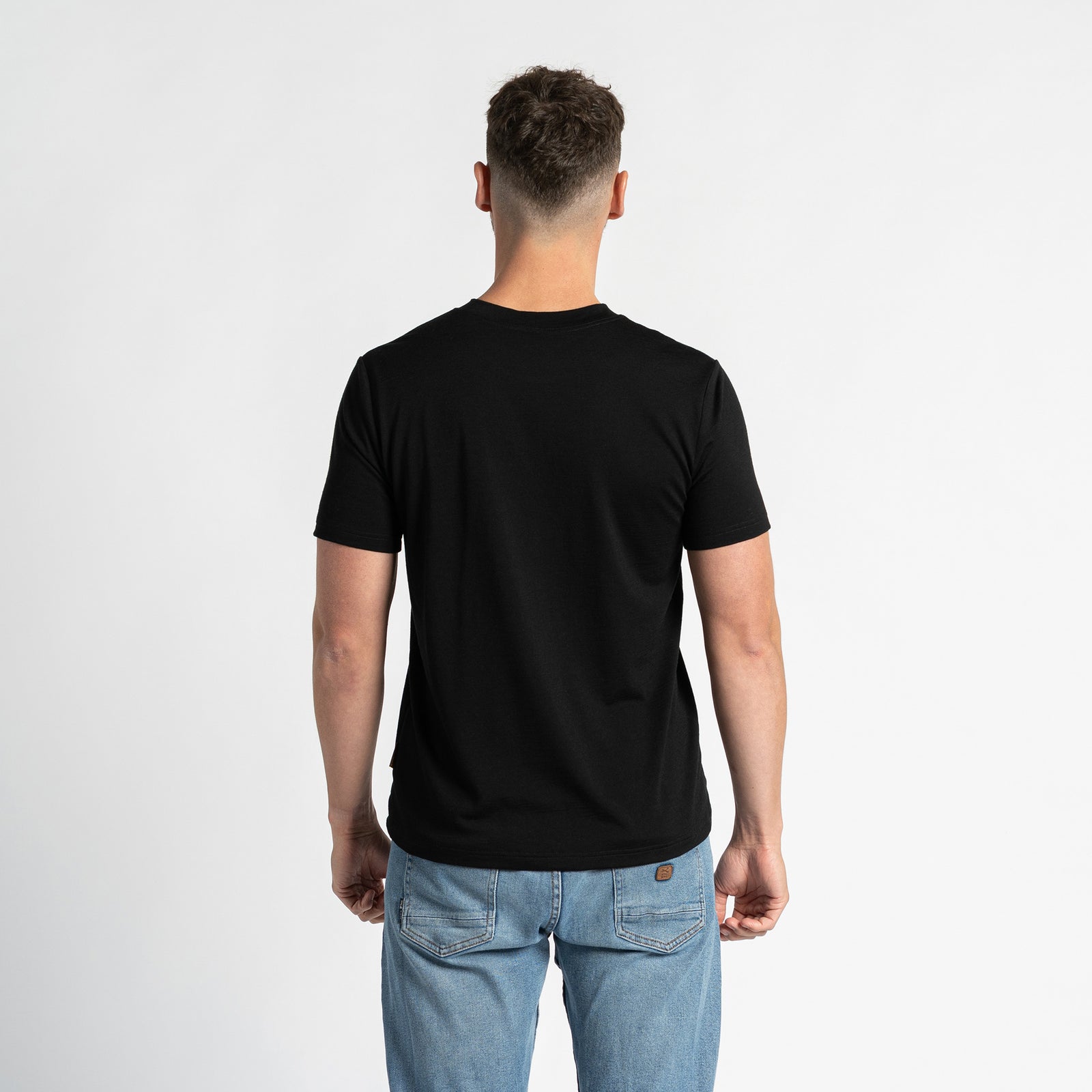 A man wearing a merino wool black t-shirt and blue jeans is shown from the chest up against a light background.  He has short brown hair and a light beard, and is looking slightly to the side. The menique black t-shirt is the primary focus, showcasing its fit and appearance.