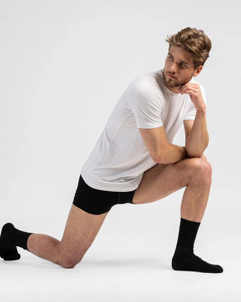 A man wearing a white t-shirt, black boxer briefs, and black crew socks kneels on one knee against a light grey background. He rests his arm on his raised knee and looks thoughtfully back over his shoulder.