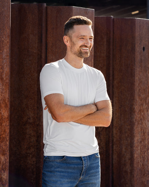 A man stands outdoors in bright sunlight with his arms crossed over his chest, smiling warmly and looking off to the side. He is wearing a plain white short-sleeved t-shirt and blue jeans, positioned in front of large, rusted steel pillars.