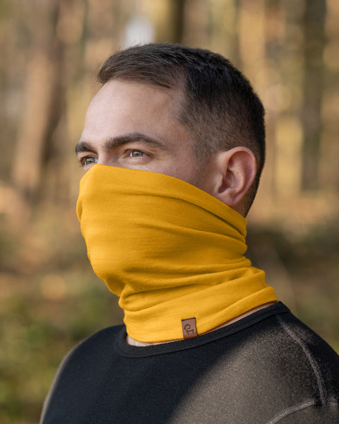 A person with short, dark hair and light eyes is shown wearing a spicy yellow neck gaiter that covers their mouth and nose. They are also wearing a black long-sleeved shirt, and the blurred background suggests an outdoor setting with trees.