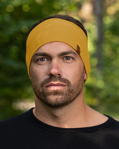A close-up of a man's face shows him wearing a spicy yellow menique headband with a small brown tag. He has short, dark hair, light eyes, and a short beard. He is wearing a black shirt and looking directly at the camera with a serious expression. The background is softly blurred green and brown, suggesting an outdoor setting.