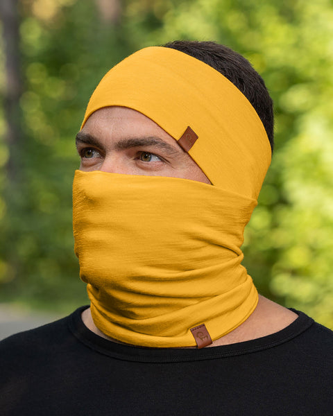 A person with short, dark hair is shown wearing a spicy yellow headband and a matching neck gaiter that is pulled up to cover their mouth and nose, with only their eyes visible. They are wearing a black long-sleeved shirt, and the blurred background suggests an outdoor setting with green foliage.
