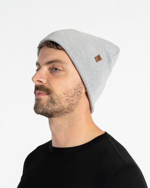 A person with light skin, short brown hair, and a beard is shown in profile against a white background. They are wearing a light gray knit beanie with a small brown tag on the side. They are also wearing a black t-shirt.