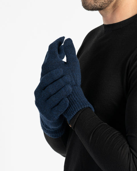 A close-up studio shot shows a person with a beard wearing blue merino wool knit gloves. The gloves have a ribbed cuff and appear to be a snug fit. The person is also wearing a long-sleeved black shirt. The background is plain white.