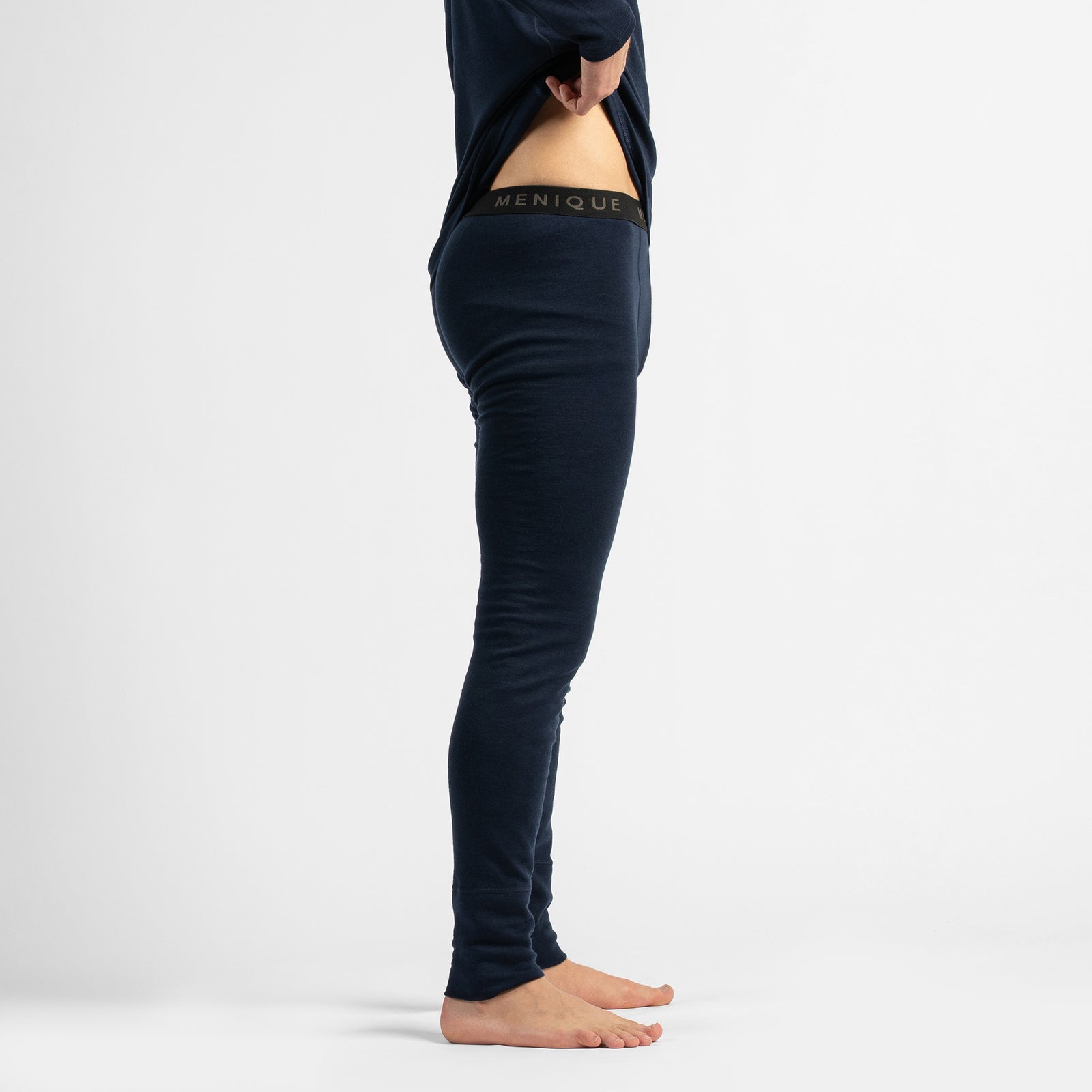 A full-length studio portrait of a barefoot man standing against a white background, wearing dark navy thermal base layers while adjusting his collar.