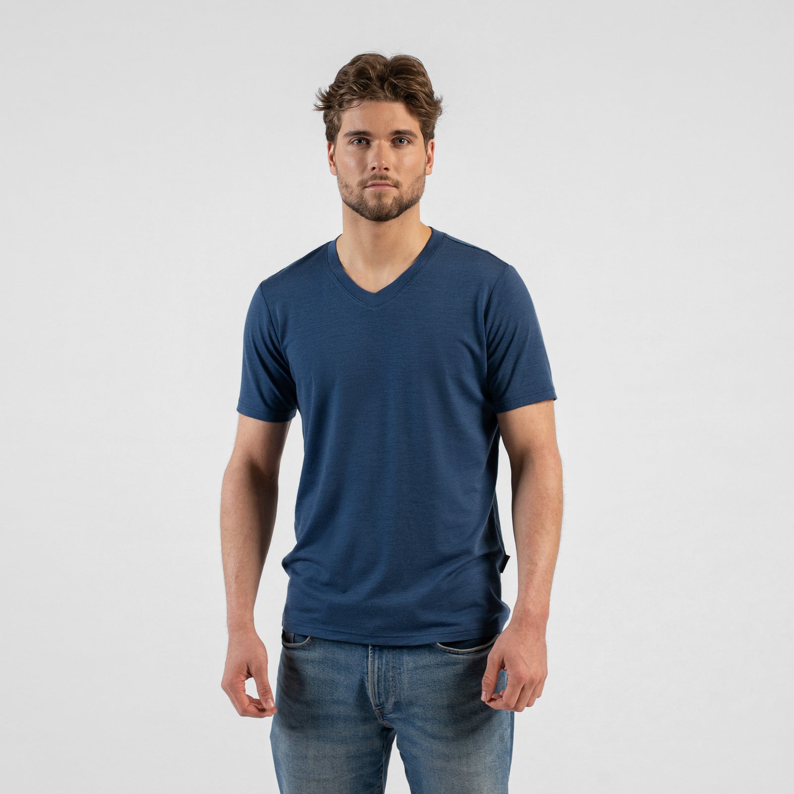 A man stands leaning casually with his right hand on a black stool, looking off to the right. He wears a denim blue v-neck t-shirt and blue jeans against a light grey background.