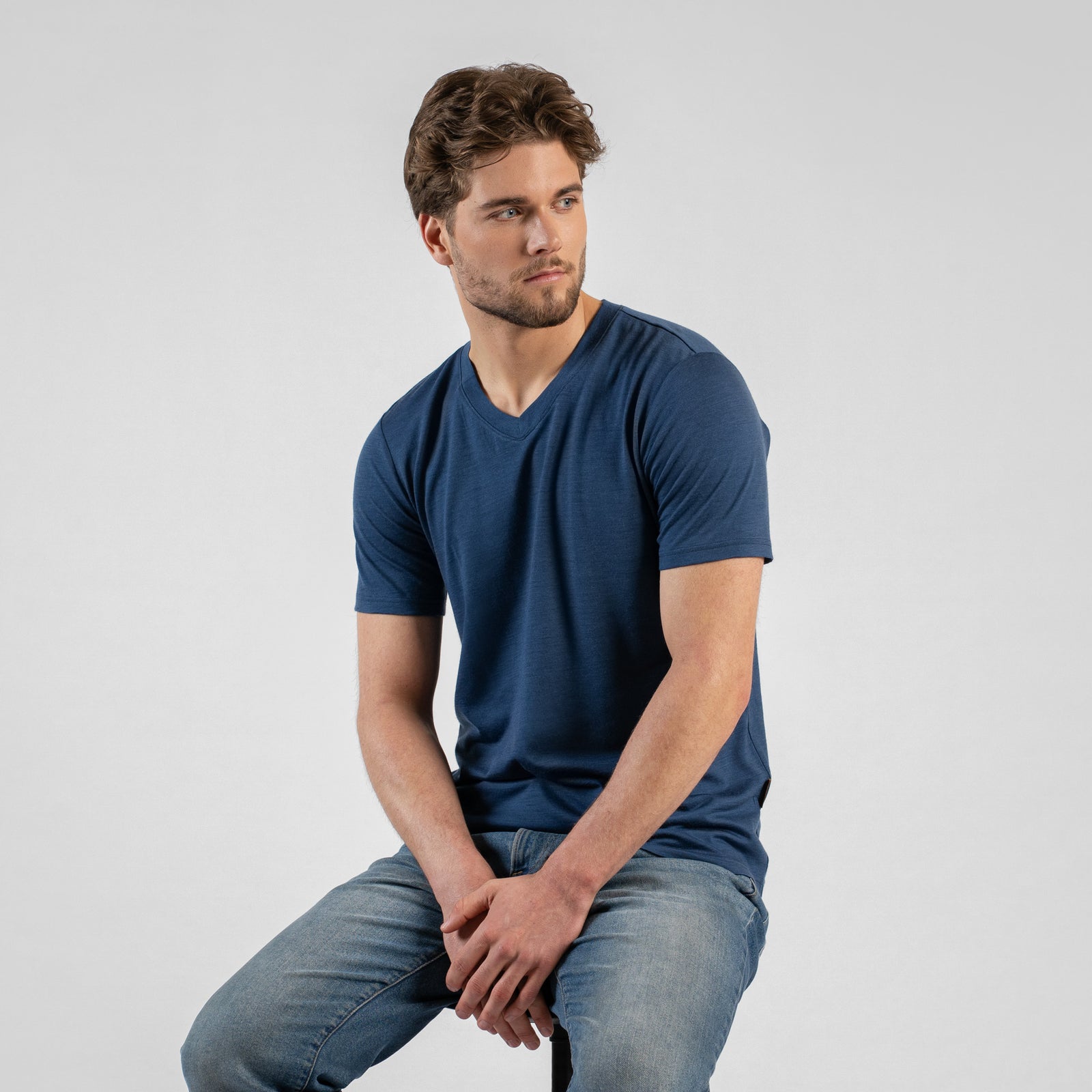 A studio portrait of a man sitting on a black stool, looking off to the right side of the frame with his hands clasped between his knees. He wears a denim blue v-neck t-shirt and blue jeans.