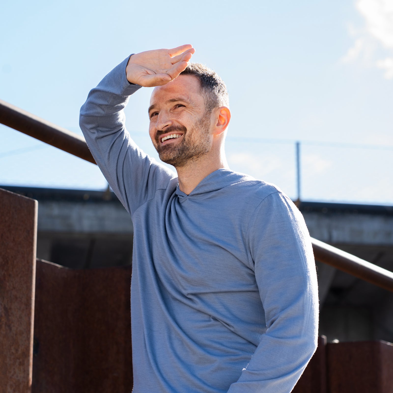 A smiling man standing outdoors near rusted metal barriers, wearing a dusty blue hoodie with the hood pulled up over a black baseball cap. He has one hand in his jeans pocket and is looking to the side.