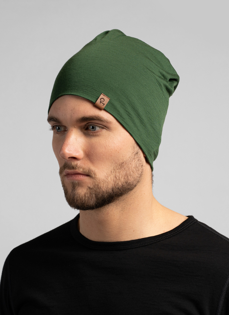 A close-up portrait shows a man wearing a snug green beanie featuring a small square leather logo tag. He is looking slightly off-camera while wearing a simple black crew-neck top against a light background.